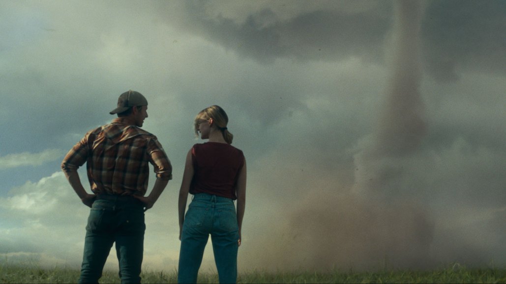 A horizontal still from the movie 'Twisters' a man and a woman stand next to each other in a field, backs to the camera, and share a look while an active tornado is nearby.